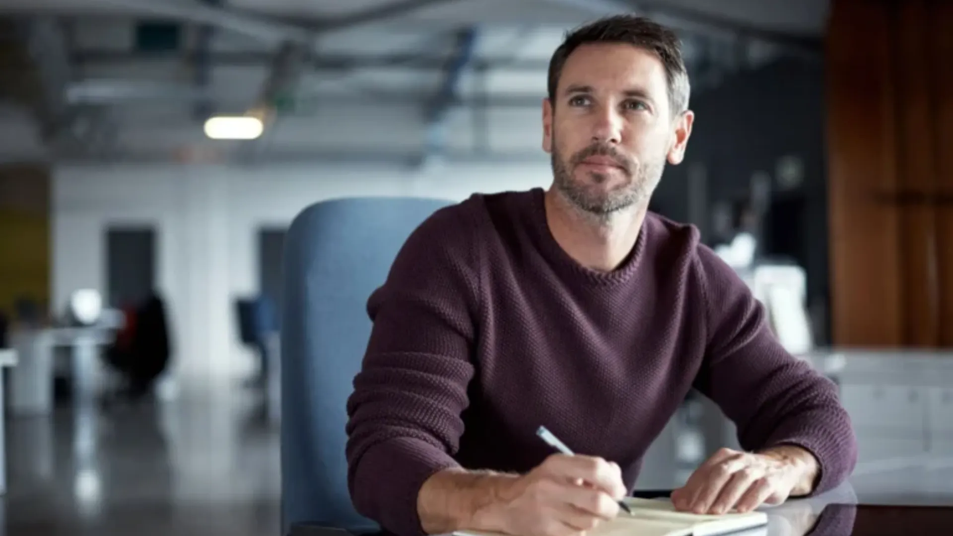 Man with beard wearing a maroon sweater, sitting and writing.