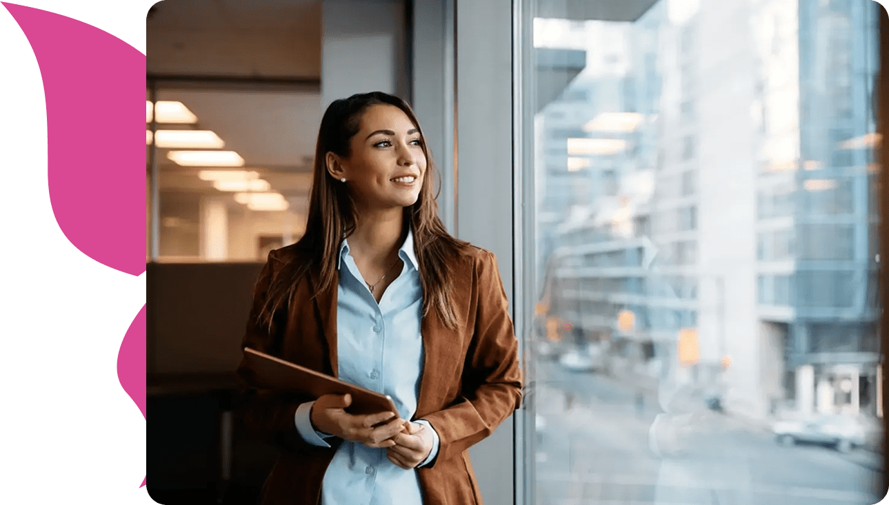 Woman in brown blazer holding tablet, looking out window with a smile.